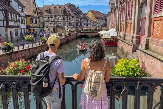 Two travelers on a bridge above the canal in Colmar with half timbered houses and a sightseeing boat